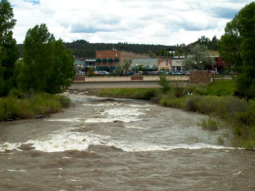A river flows through a green landscape with a small town and buildings visible in the background under a cloudy sky.