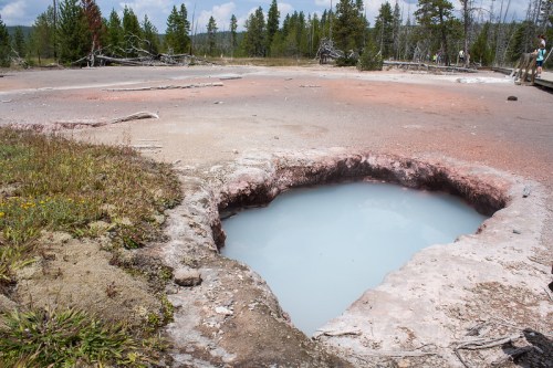 A geothermal hot spring with pale blue water surrounded by rocky terrain and sparse vegetation in a natural landscape.