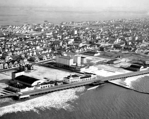 Aerial view of a coastal town featuring a pier, buildings, and a beach, captured in black and white.