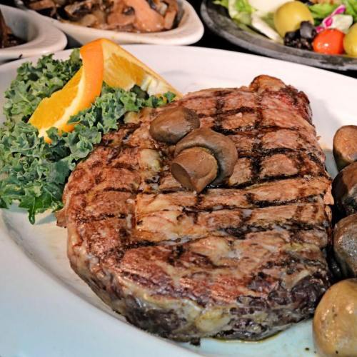 A grilled steak with mushrooms, garnished with kale and an orange slice, served on a white plate.