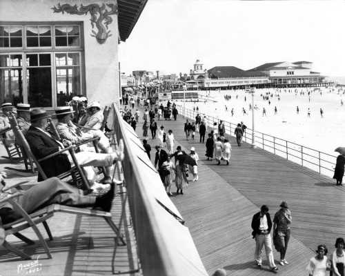 Black and white photo of a busy beach boardwalk with people walking, lounging, and enjoying the seaside atmosphere.