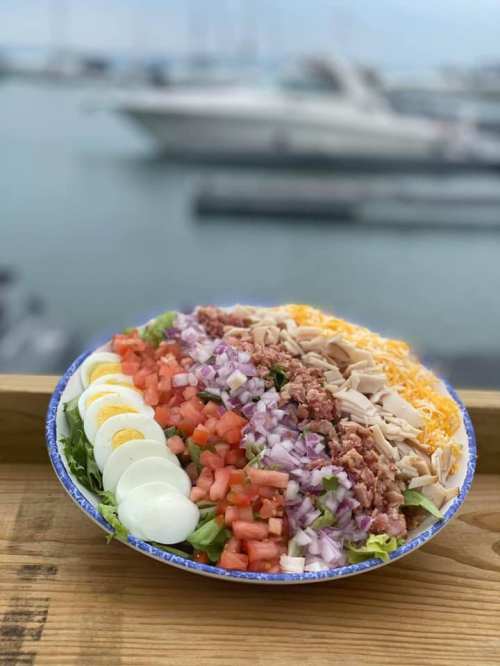A colorful salad with layers of lettuce, tomatoes, onions, cheese, chicken, and sliced eggs, set against a marina backdrop.