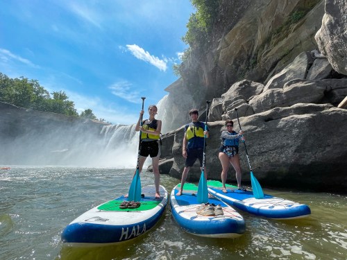 Three people stand on paddleboards in a river near a waterfall, surrounded by rocky cliffs and greenery.