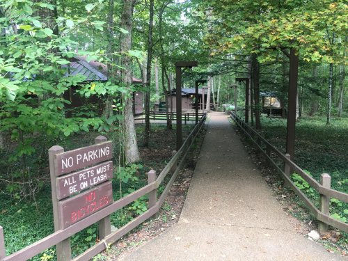 A wooded path leads to cabins, with a sign stating "No Parking" and "All pets must be on leash."