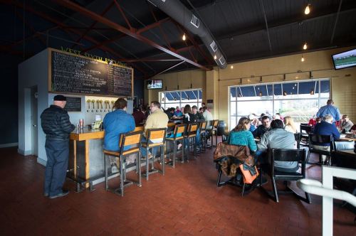 A lively bar scene with patrons seated at a wooden counter and tables, enjoying drinks in a spacious, well-lit venue.