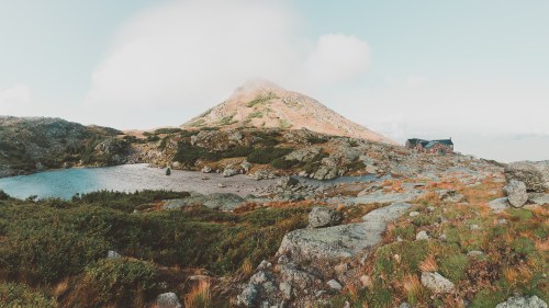 A serene mountain landscape featuring a peak, a small lake, and a cabin surrounded by rocky terrain and greenery.
