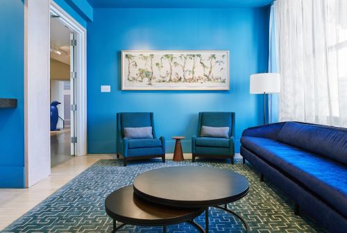 A bright blue room featuring a blue sofa, two chairs, a round coffee table, and a large framed artwork on the wall.
