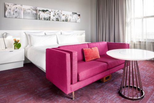A modern hotel room featuring a large bed, a pink sofa, and a round table, with artwork of sheep on the wall.