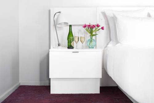 A modern bedside table with a lamp, a bottle of wine, glasses, and a vase of flowers next to a neatly made bed.