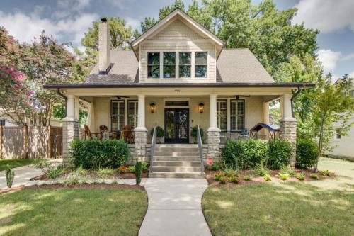 A charming two-story house with a front porch, surrounded by greenery and colorful flowers under a cloudy sky.