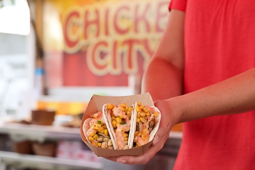 A person holds a tray with three colorful tacos filled with chicken, corn, and toppings, in front of a food stall.