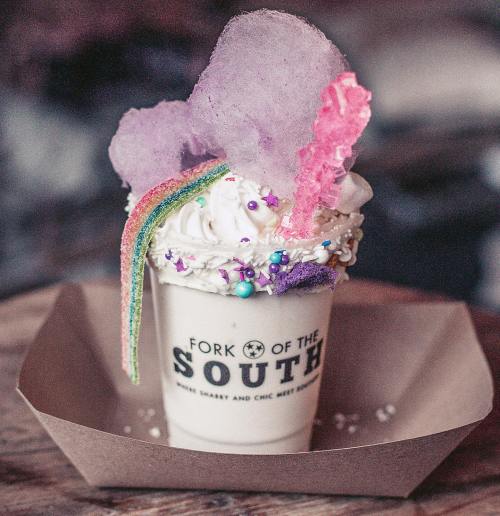 A whimsical dessert in a cup topped with whipped cream, cotton candy, rainbow sprinkles, and colorful decorations.