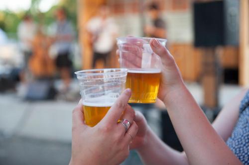 Two hands clink plastic cups of beer in a lively outdoor setting with a band performing in the background.