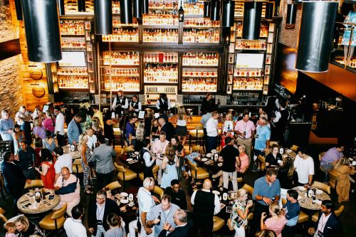 A bustling bar scene with a large crowd, shelves of liquor, and people socializing at tables and the bar.
