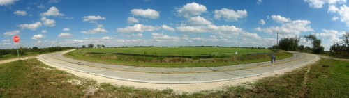 A panoramic view of a rural road intersecting with a stop sign, surrounded by green fields and a blue sky with clouds.