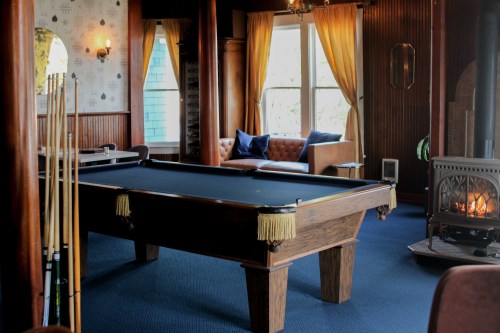 A cozy room featuring a pool table, a fireplace, and a comfortable seating area with large windows and warm lighting.