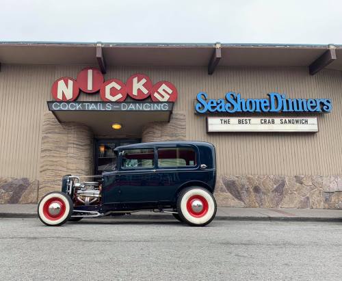 A vintage car parked outside a restaurant with a sign for crab sandwiches and a retro-style cocktail bar.