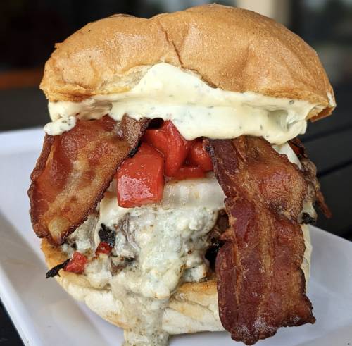 A close-up of a juicy burger topped with bacon, roasted red peppers, and creamy sauce, served in a soft bun.