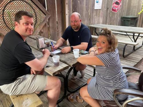 Three people sitting at a wooden table outdoors, smiling and engaged in conversation, with drinks in front of them.