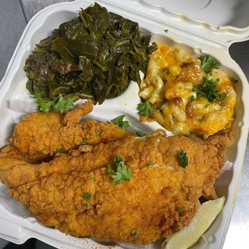 A takeout container with two fried fish fillets, collard greens, and cheesy macaroni, garnished with parsley.