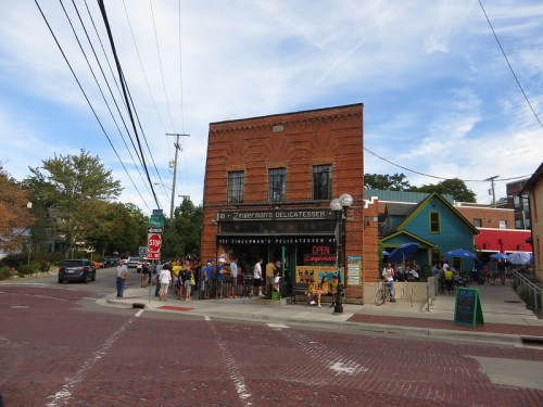 A brick building with "Zingerman's Delicatessen" sign, bustling with people outside on a sunny day.