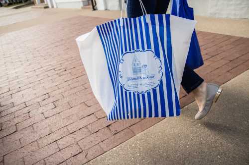 A person walks on a brick path, carrying a large blue and white striped shopping bag with a logo.