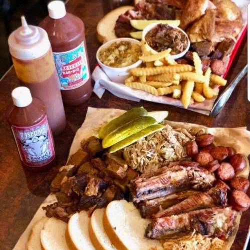 A spread of barbecue dishes including ribs, pulled pork, fries, cornbread, pickles, and sauces on a wooden table.
