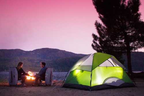 A couple sits by a campfire near a tent, with a scenic lake and mountains in the background at sunset.