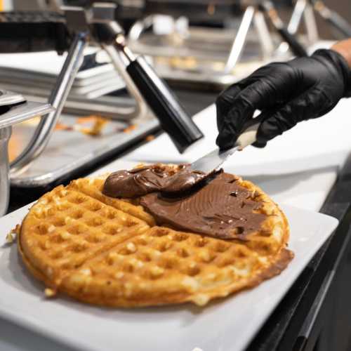 A hand spreads chocolate spread on a freshly made waffle on a white plate at a food station.