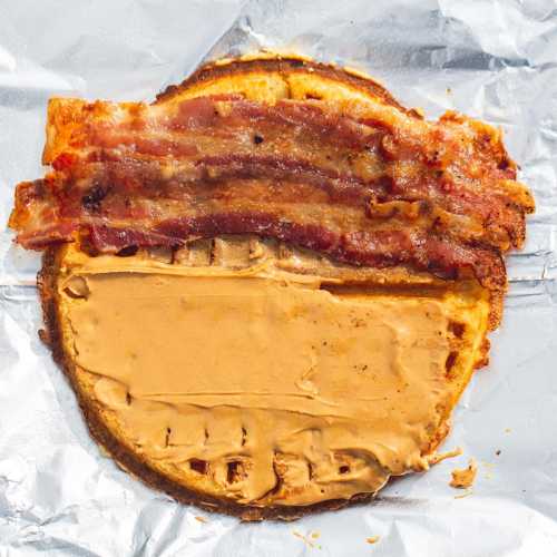 A toasted waffle topped with peanut butter and crispy bacon strips, wrapped in foil.