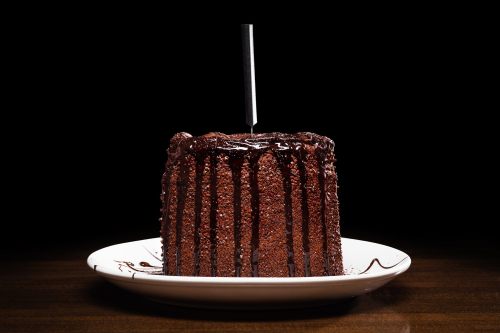 A rich chocolate cake with drizzled chocolate sauce, presented on a white plate against a dark background.