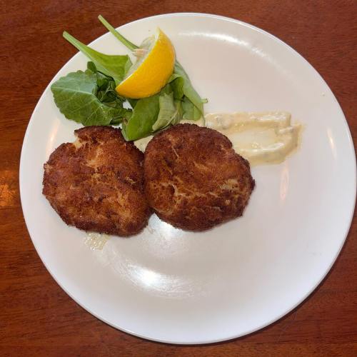 Two golden-brown crab cakes on a white plate, accompanied by a lemon wedge, greens, and a dollop of sauce.