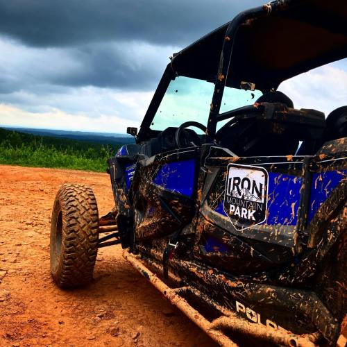 A muddy off-road vehicle parked on a dirt trail with a scenic view of mountains and cloudy skies in the background.