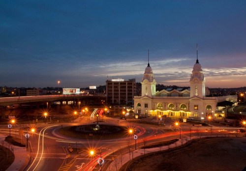 A historic building illuminated at dusk, surrounded by a circular road and light trails from passing vehicles.