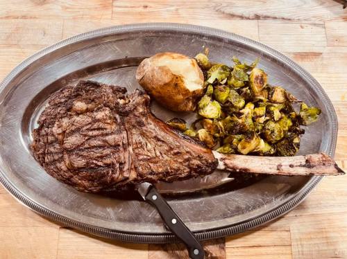 A large ribeye steak on a silver platter, accompanied by a baked potato and roasted Brussels sprouts.