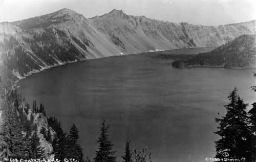 Black and white photo of Crater Lake, surrounded by mountains and trees, showcasing its calm waters.