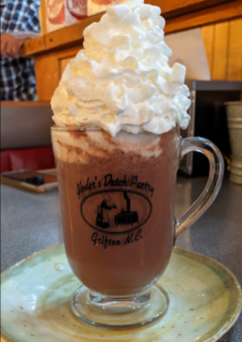 A glass mug filled with hot chocolate topped with a generous swirl of whipped cream, sitting on a plate.