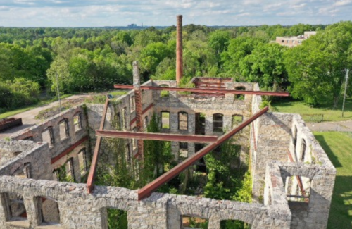 Aerial view of a stone ruin with overgrown vegetation and a tall chimney, surrounded by lush green trees.