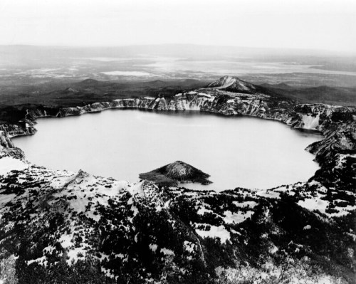 Aerial view of a large volcanic crater lake surrounded by rugged terrain and distant mountains, in black and white.