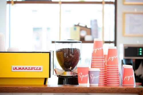 A coffee shop counter featuring a yellow La Marzocco espresso machine, a bean grinder, and stacked colorful cups.