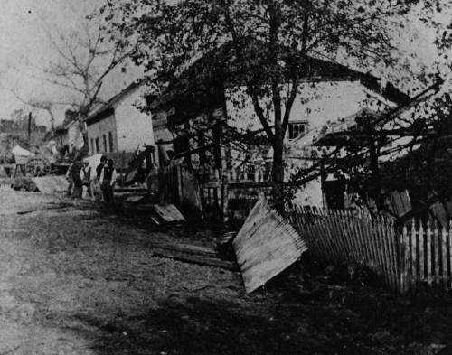 Historic black-and-white photo of a damaged building with people and debris in a rural setting.