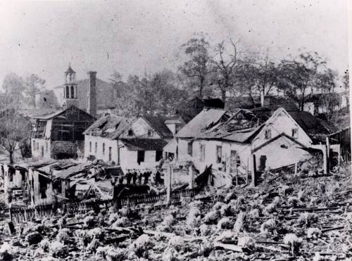 Black and white photo of damaged buildings and trees, depicting destruction in a rural area.
