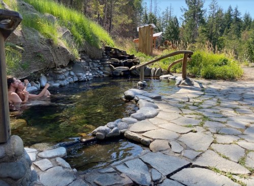 A serene outdoor hot spring surrounded by greenery, with a person relaxing in the water and a stone pathway nearby.