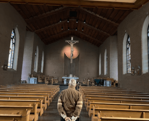 A person stands in a church, facing the altar with a crucifix, surrounded by empty wooden pews and stained glass windows.