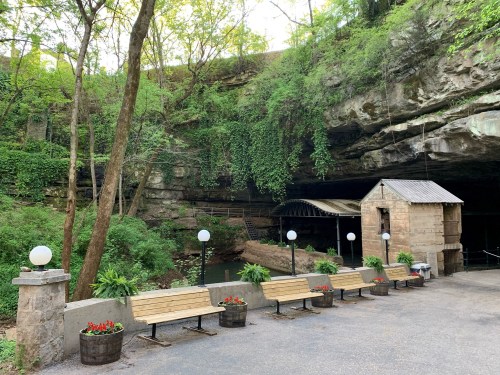 A serene outdoor scene featuring benches, planters, and a small building near a rocky cave entrance surrounded by greenery.