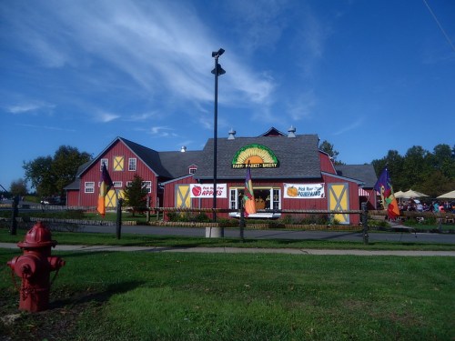 A red barn-style market with colorful banners, surrounded by green grass and a blue sky.
