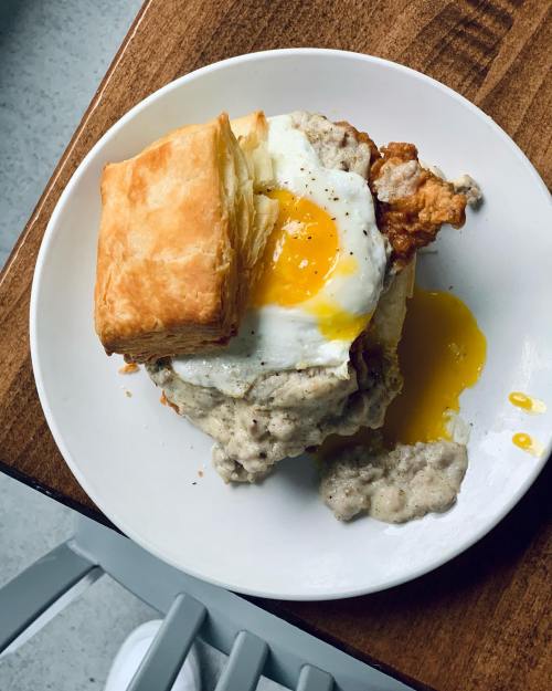 A plate with a flaky biscuit topped with fried chicken, a sunny-side-up egg, and creamy sausage gravy.