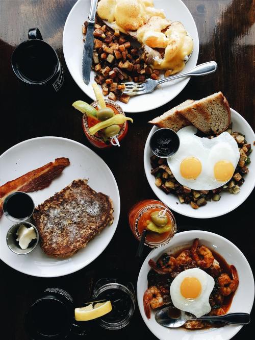 A table spread with breakfast dishes: eggs benedict, fried eggs, French toast, and drinks, all on white plates.