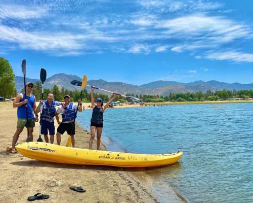 Four people stand by a yellow kayak on a beach, smiling and enjoying a sunny day by the water with mountains in the background.