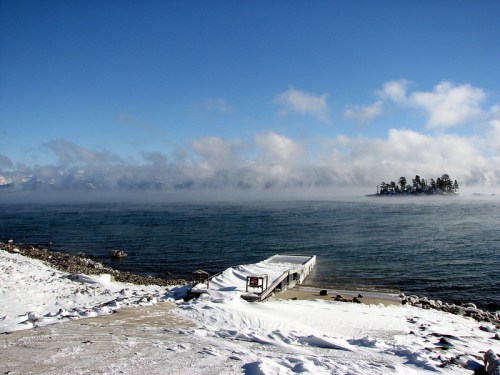 A snowy shoreline with a dock leading to a misty lake and an island in the distance under a clear blue sky.